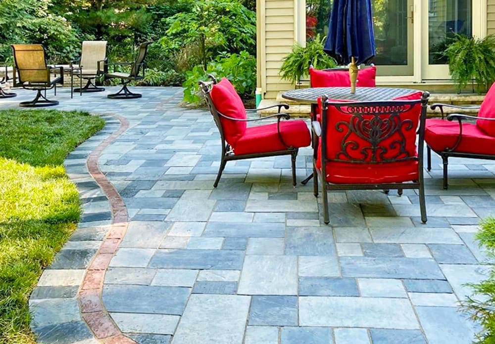 Outdoor patio with stone pavers, red chairs, and a table, surrounded by greenery.