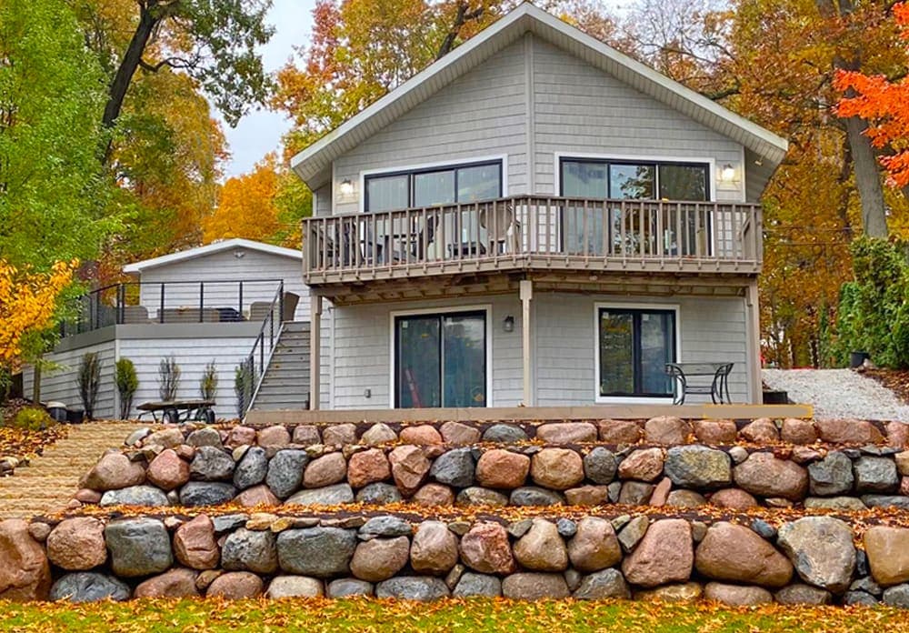Modern gray house with balcony, surrounded by autumn foliage and stone landscaping.
