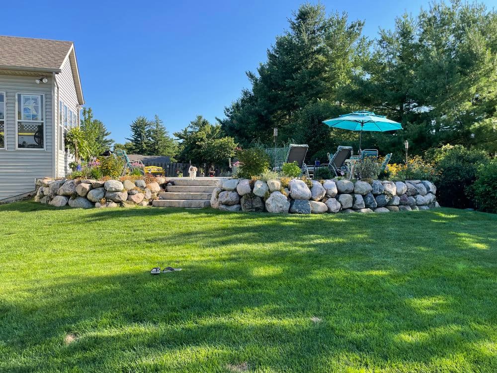 Lush backyard with stone retaining wall, greenery, seating area, and umbrella on a sunny day.