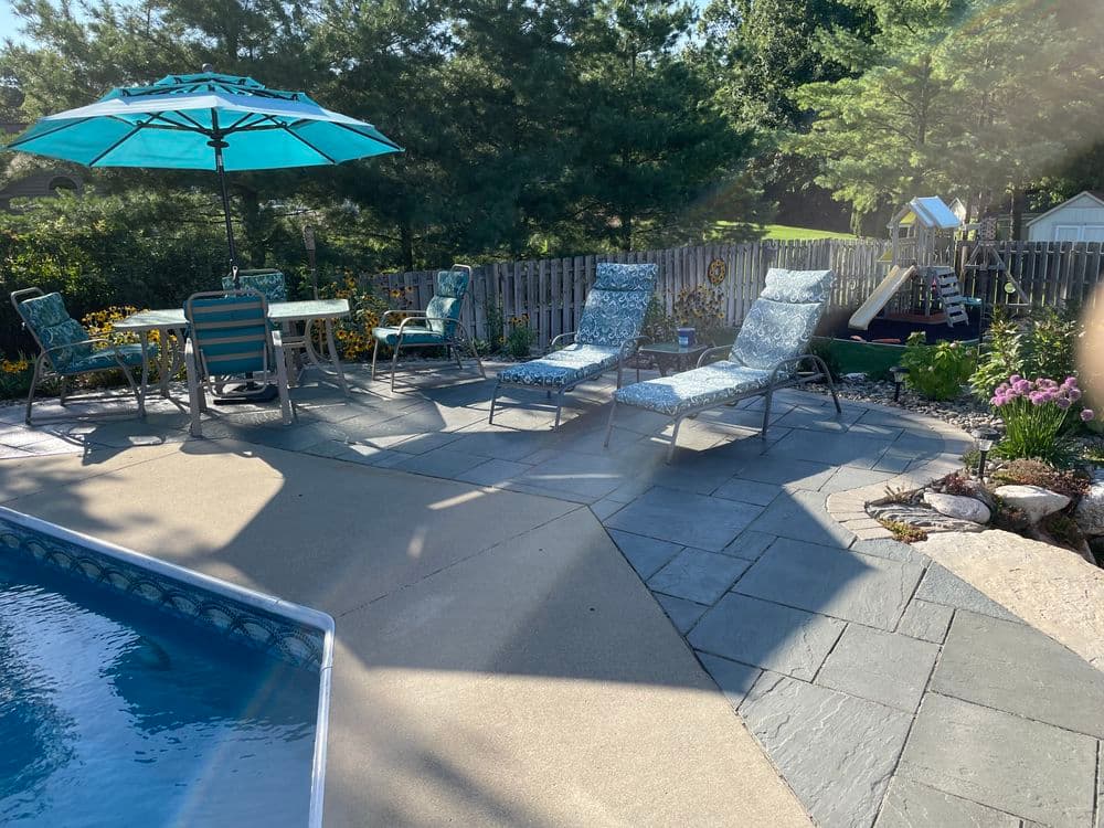 Poolside patio with lounge chairs, table, umbrella, and lush garden in sunny backyard setting.